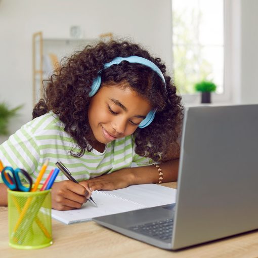 Happy school child sitting at desk with newly donated laptop by Give IT Forward, having online class and writing in notebook